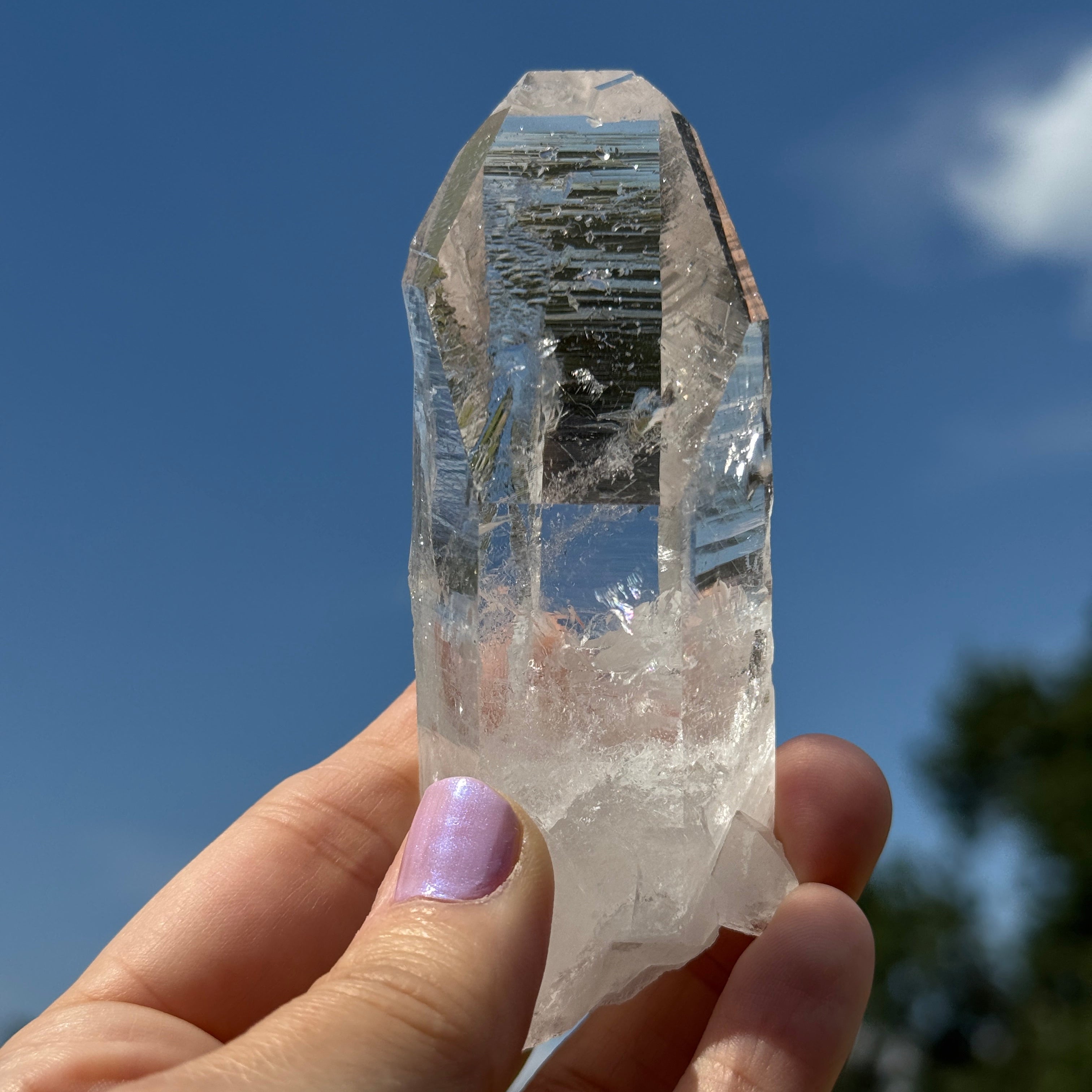 A person's hand holding a clear quartz crystal point against a bright blue sky background.