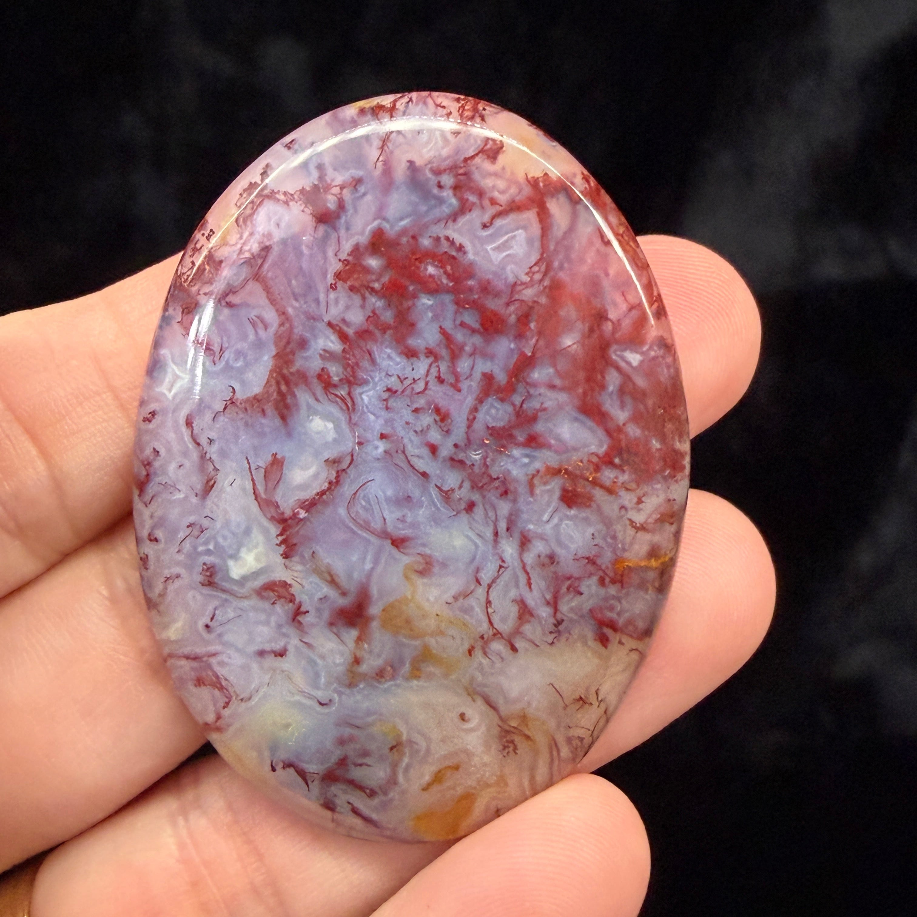 Hand holding a Moss Agate Cabochone with red, and white colors against a dark background